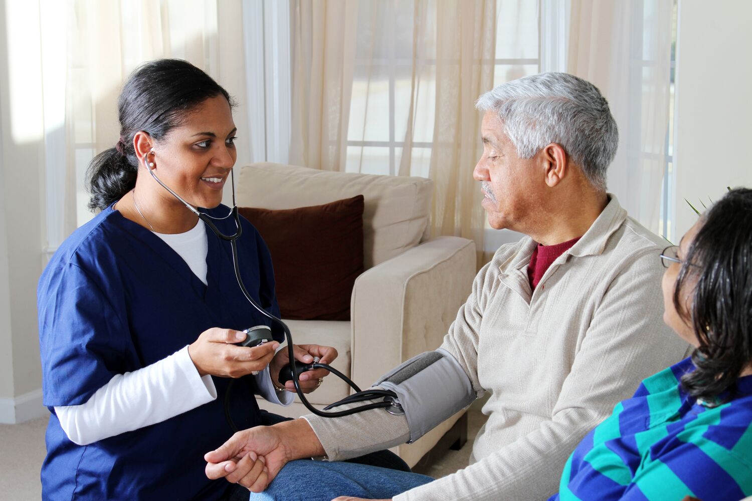 A home health care worker smiles as she checks the blood pressure of an elderly man sitting on a couch as his spouse looks on (BigStock 13926641)