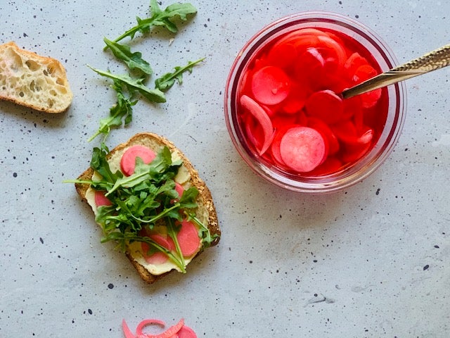 Jar of pickled radishes and onions beside a piece of whole wheat toast with hummus, arugula and pickled radishes